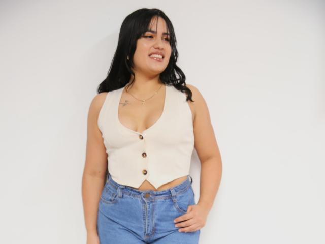 TifaniGreys met zwart haar, die een witte vest zonder mouwen draagt met knopen, waarbij haar borsten zichtbaar zijn, en blauwe jeans, lachend, staand tegen een witte muur..