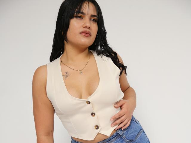 TifaniGreys has long black hair, light brown skin, and a small tattoo on her left chest. She wears a sleeveless, cream-colored, button-up top with brown buttons, revealing cleavage. She has a confident, slightly relaxed posture with her right hand on her hip. She wears blue jeans and a simple necklace. The image is a close-up shot with a plain white background. The composition emphasizes her upper body and facial expression.