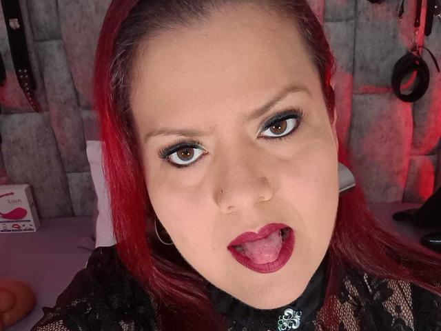 VanesaMoka mit roten Haaren, offenem Mund, herausgesteckter Zunge, trägt eine schwarze Spitzenbluse, Reifenohrringe und dunkles Make-up, blickt mit braunen Augen nach oben und befindet sich in einer sexy, provokanten Pose..