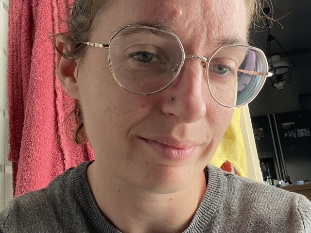 JolieJoelle has freckles, a nose piercing, and wears round glasses with a slight reflection. She's in a gray sweater, with a pink towel behind her.