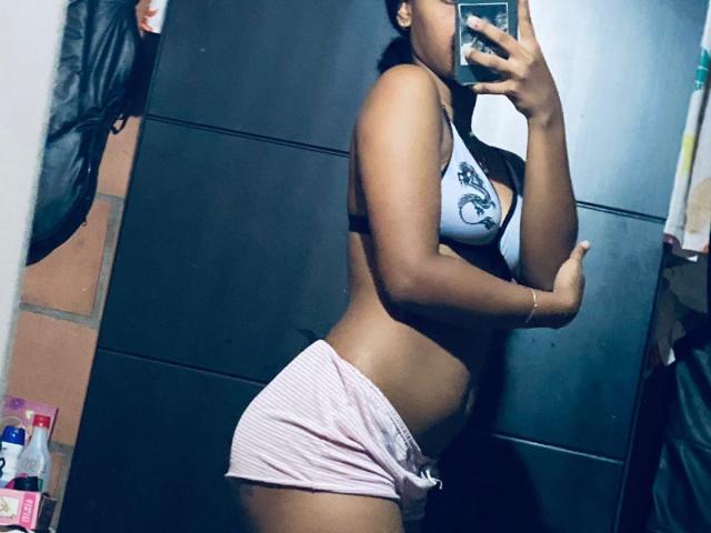 HottieLoveTime with dark skin, small tits in white bra, pink shorts, holding phone in mirror selfie, showing side profile and belly, in dimly lit room.
