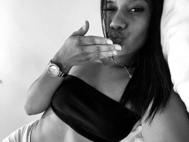 Black and white photo of HottieLoveTime with dark straight hair, nose ring, and wristwatch, kissing her fingers, wearing a strapless black top, showing her small tits.