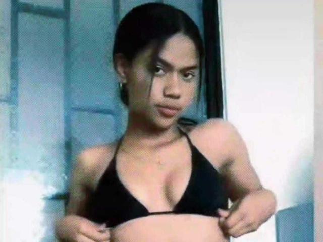 HottieLoveTime with dark hair, wearing a black bikini top, showing off her small, perky tits, looking down with a serious expression, standing in a slightly blurry photo.