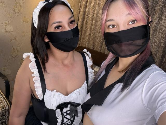 Two SexLoveDuo in French maid outfits, wearing black masks, one with pink highlights in her hair. One has big boobs in a corset, the other has a white shirt.