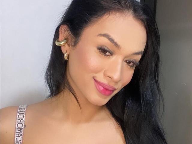 Lia, con su largo cabello negro, aretes dorados, lápiz labial rosa y una sonrisa, muestra sus senos en un vestido brillante con tirantes. Sus ojos marrones están ligeramente bajos..