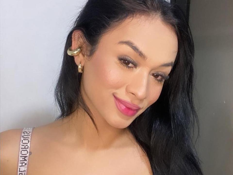 Lia, com seus longos cabelos pretos, brincos de ouro, batom rosa e um sorriso, exibe seus seios em um vestido brilhante com alças finas. Seus olhos castanhos estão levemente voltados para baixo..