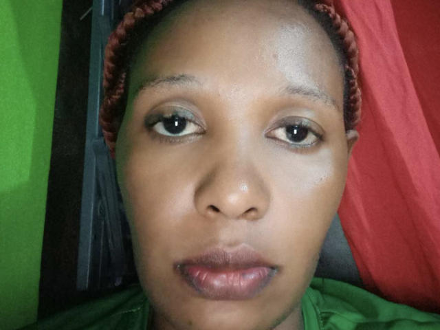 NkiiOne's face, with brown skin, small nose, and brown eyes, wearing a red braided headscarf, green shirt, and red background, showing slightly shiny skin and neutral expression.