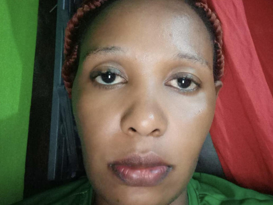 NkiiOne's face, with brown skin, small nose, and brown eyes, wearing a red braided headscarf, green shirt, and red background, showing slightly shiny skin and neutral expression.