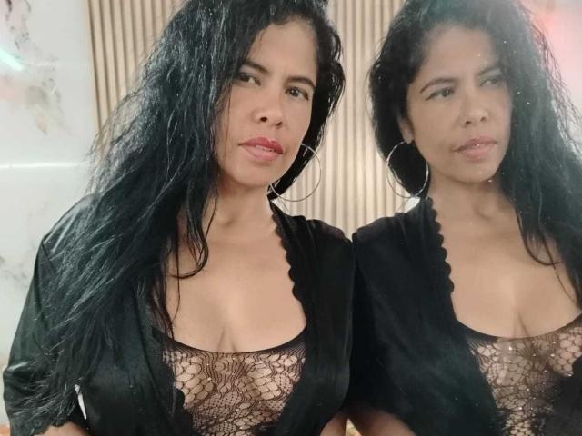 Sofia Lustery, com longos cabelos pretos, vestindo uma blusa preta de renda que mostra seus seios e um robe preto, em pé lado a lado, olhando para a câmera com batom vermelho..