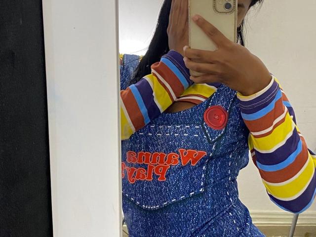 HottieLoveTime in a blue, textured dress with red button, striped yellow, purple, and red sleeves, holding a phone, mirror selfie, showing brown skin, long dark hair.