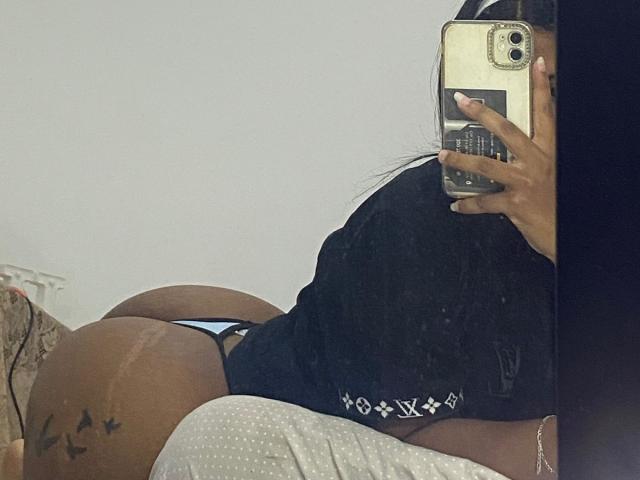 HottieLoveTime's butt in blue thong, wearing black shirt, lying on bed, holding phone, dark skin, tattoo of birds on right thigh, reflection in mirror, white wall.