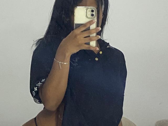 HottieLoveTime with dark skin, long black hair, wearing a black polo shirt, holding a phone covering her face, showing her small tits and black thong. Mirror selfie.