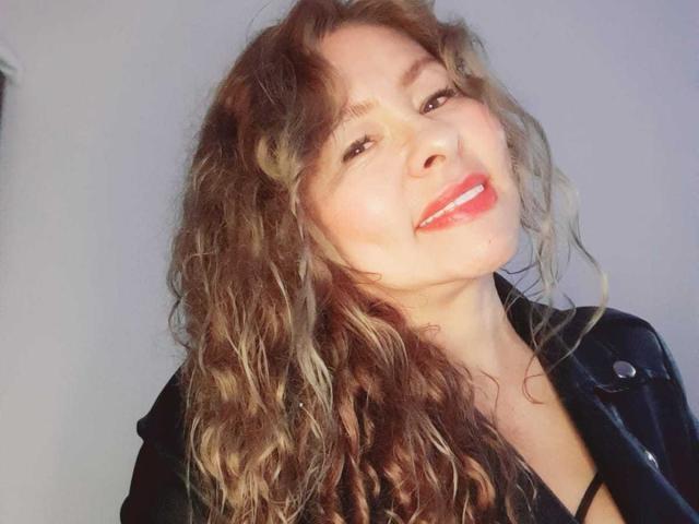 MiaAmoretti with wavy brown hair, red lipstick, and a black leather jacket, smiling slightly. She has a cute face and a small nose. Her tits are not visible.