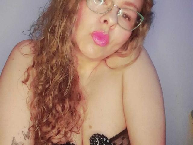 MiaAmoretti with curly brown hair, pink lipstick, glasses, and a black polka-dot bra, pouting her pink lips, showing her bare tits, and a tattoo on her right arm.