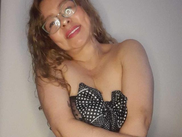 MiaAmoretti with curly brown hair, glasses, and pink lipstick, smiling, arms crossed, wearing a black polka dot strapless top, showing her boobs.