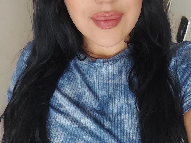 LauraRichis with black wavy hair, wearing a blue tie-dye rib-knit shirt, showing her pouty pinkish-brown lips, and her small, cute, round ass barely visible.