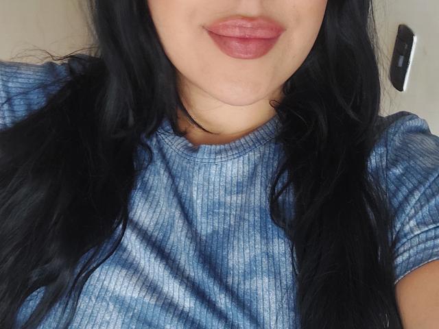 LauraRichis with long black hair, wearing a blue tie-dye shirt, has pinkish lipstick on her pouty lips, showing a slight double chin, and a small mole on her neck.