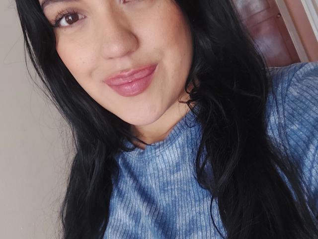 LauraRichis with long black hair, pink glossy lips, and a slight smile, wearing a blue tie-dye rib-knit sweater, brown eyes, and a light skin tone.