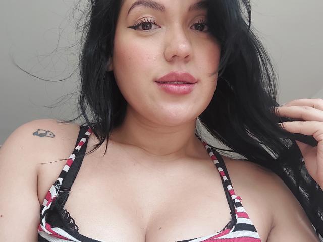 LauraRichis with black hair, wearing a striped top, showing her tits, has a small tattoo on her left shoulder, and is touching her hair with a slight smirk.