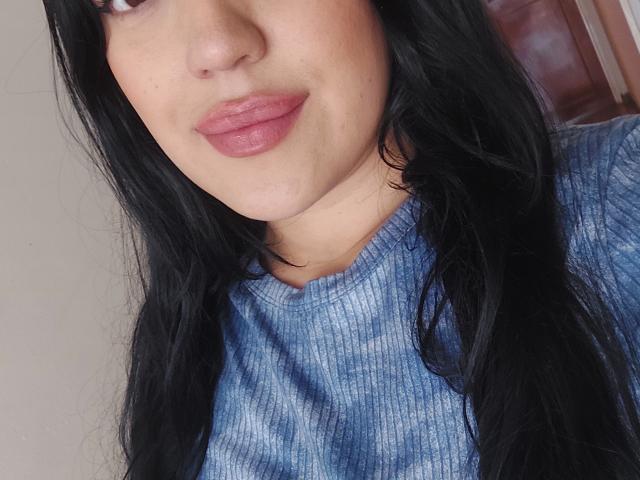 LauraRichis with long black hair, pinkish lips, and a blue tie-dye shirt, showing a subtle smile, has a small chin cleft and a slightly freckled face.