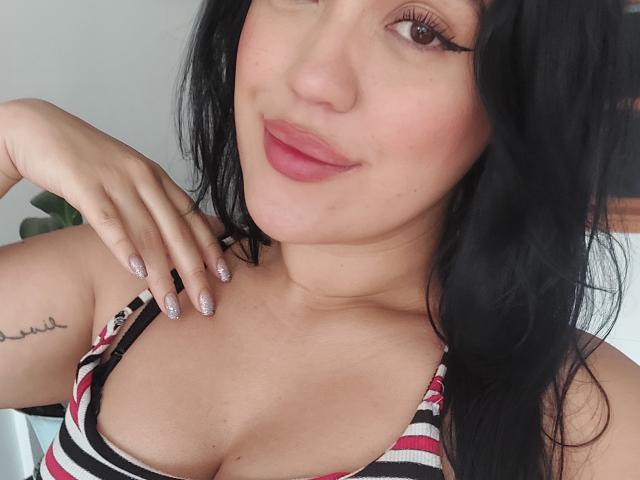 LauraRichis has black hair, pink lips, and sparkly nails, wearing a striped top with cleavage, showing a small tattoo on her right shoulder, smiling.