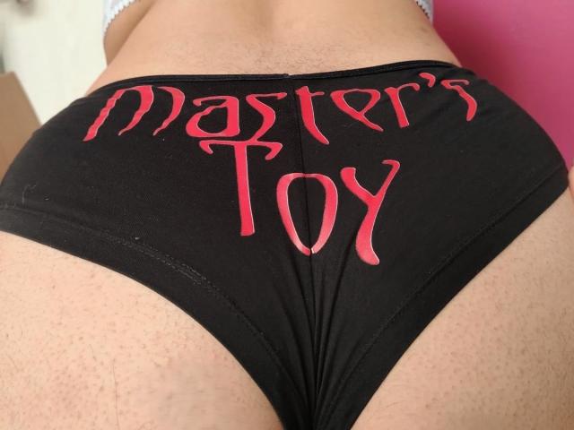 Les culottes noires de TifaniGreys avec le texte rouge "Master's Toy", mettant en valeur ses fesses galbées avec une texture de peau visible et une ceinture blanche..