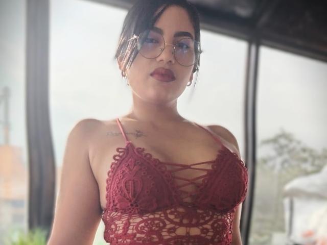 TifaniGreys with dark hair in glasses, wearing a red lace bralette that shows her tits, standing in front of a window with sunlight. Tattoos on her chest.