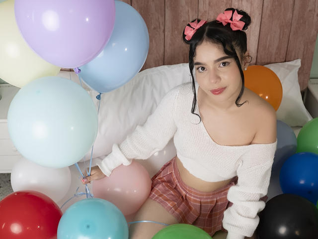 LissyCarter with black hair in pigtails, pink bows, white off-shoulder sweater, pink plaid skirt, surrounded by colorful balloons, sitting on bed. Boobs partially visible.