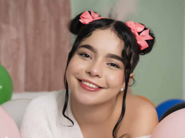 LissyCarter has black hair in double buns with pink bows, a cute smile, and is wearing a white top, showing her bare shoulders and a pearl earring.