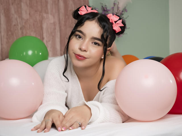 LissyCarter with pink bows in black hair, lying on white table, surrounded by balloons, wearing off-shoulder white sweater, smiling, pink nails, soft makeup, cute, sexy.