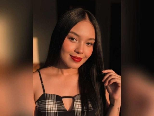MissOrtizz with long black hair, red lipstick, smiling, wearing a black and white plaid dress with a keyhole cutout, holding her hair, soft sunlight.