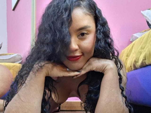 MissOrtizz with curly black hair, red lipstick, and hands under her chin, smiling at the camera, her bare tits slightly visible, leaning forward.