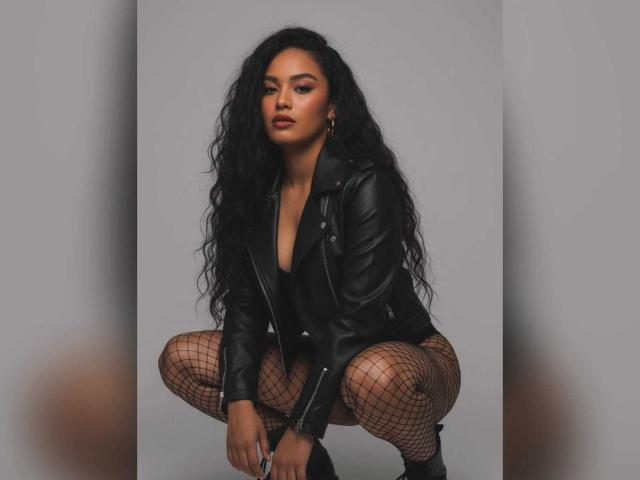 MissOrtizz is squatting, wearing a black leather jacket, fishnet stockings, and black boots. She has long, curly black hair, dark makeup, and a serious expression. Boobs are visible.
