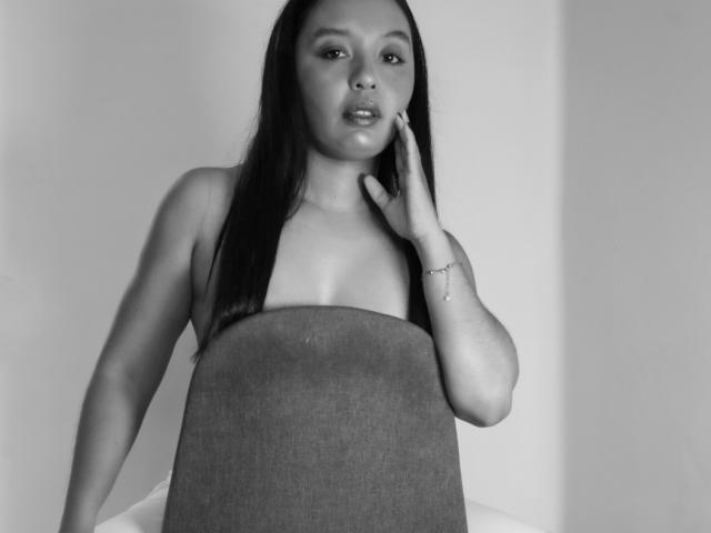 MissOrtizz with long black hair, wearing a strapless top, touching her cheek with one hand, showing her small tits and a slim, sexy body, in a black-and-white photo.