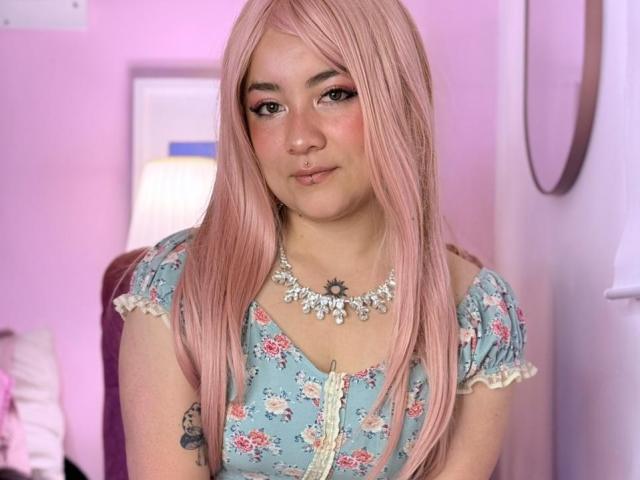 Froditaa, con i capelli rosa, indossa un abito blu a fiori e una collana di diamanti, ha un piccolo tatuaggio sul braccio destro e un delicato anello al naso, appare carina e rilassata..