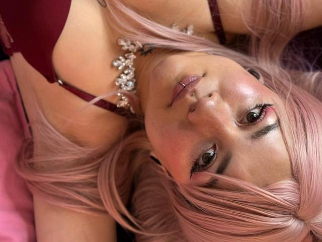 Froditaa, con i capelli rosa, reggiseno rosso e piercing al naso, sdraiata, che guarda la fotocamera, indossa una collana con gioielli, con un trucco leggero e un viso leggermente arrossato..