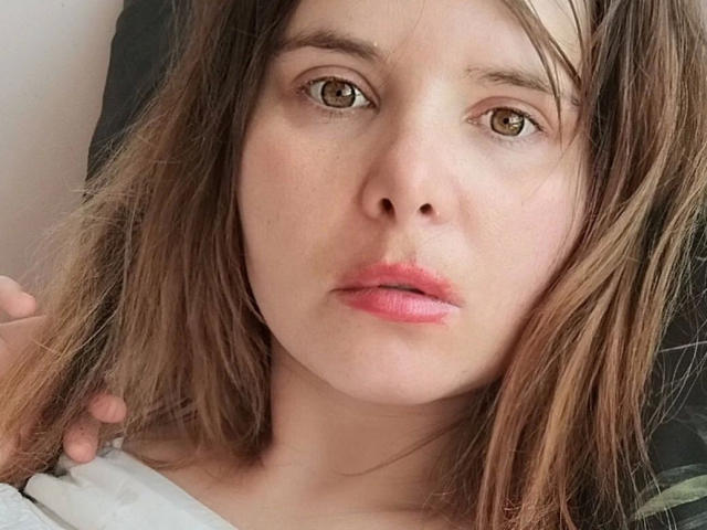 ElenaSchatz-hot has light brown eyes, pink lips, and messy brown hair, looking serious with a slight pout. Her hand touches her hair on the left.