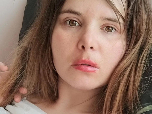 ElenaSchatz-hot has brown eyes, slightly parted pink lips, and messy brown hair. Her face shows a neutral, slightly sad expression. Her hand rests near her head.