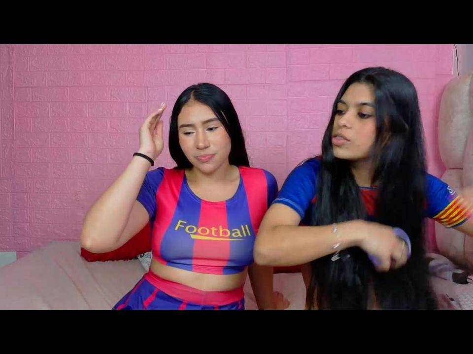 HarleyDiamondIvonne with long black hair, in a red and blue football-themed crop top, letting another HarleyDiamondIvonne with long black hair brush her hair. Pink wall in background.