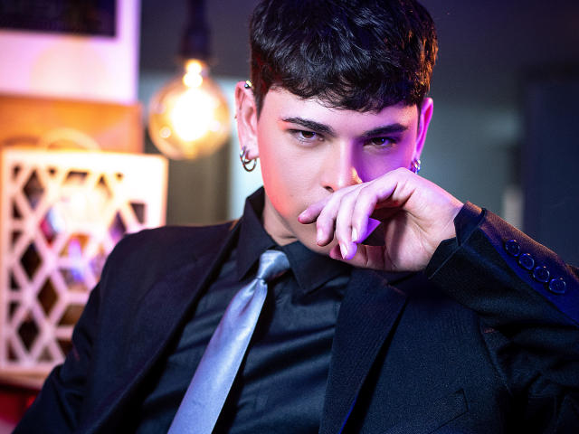 LukeHowlandy, un homme aux cheveux foncés, portant un costume noir, une cravate brillante, une chemise noire, avec des piercings aux oreilles, éclairage violet et rose, une main près de la bouche, une expression sérieuse, un ampoule suspendue en arrière-plan..