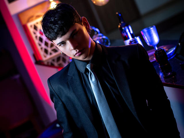 LukeHowlandy with dark hair, wearing a black suit and silver tie, leaning against a bar with a bottle, glass, and red lighting, looking serious and slightly seductive.