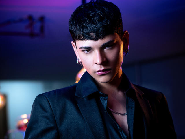 LukeHowlandy has dark hair, a serious look, and wears a black blazer over a black shirt with a silver necklace. Blue and purple lights highlight his face.