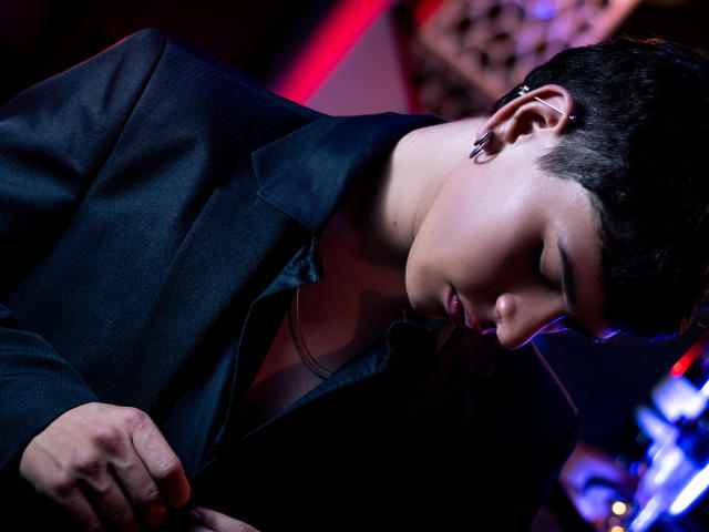 LukeHowlandy, with short black hair and ear clip, leans forward in a black suit, eyes closed, hands near chest, illuminated by purple and red lights.