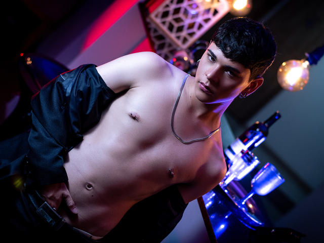 LukeHowlandy with a shirtless, toned, pale-skinned body, black jacket off, silver necklace, nipple piercings, and short dark hair, looking at the camera in a dimly lit room.