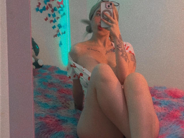 SammyViolet, con cabello rubio, blusa roja con forma de corazón y hombros descubiertos, tatuajes, sosteniendo un teléfono en la mano, sentada en una cama esponjosa azul y rosa, con las rodillas levantadas, mostrando su trasero desnudo y sus pequeños pechos..
