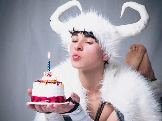 LukeHowlandy, with white furry horns and a fluffy white robe, puffs out his cheeks while blowing on a candle on a cake with red and yellow icing.