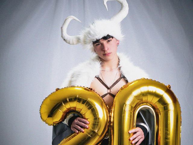 LukeHowlandy with white furry hat, horns, holding golden "20" balloons, wearing fur coat, exposed tits, brown harness, earrings, and serious expression.