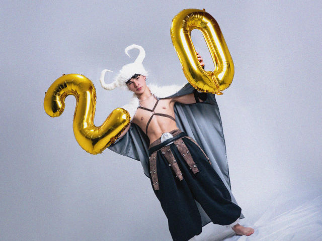 LukeHowlandy with white horned hat, holding gold "20" balloons, wearing black strappy top, loose black pants with brown belt, bare feet, and gray cape. LukeHowlandy is shirtless.