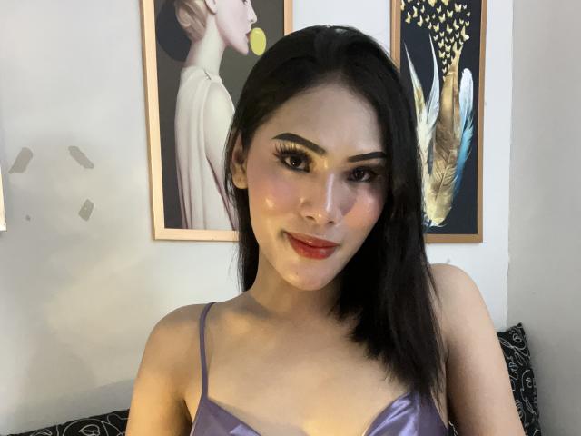 SofiaCumness with shiny face, dark eyebrows, red lipstick, and purple satin bra, smiling at camera, black hair, Asian, subtle makeup, bedroom setting, two framed portraits behind.
