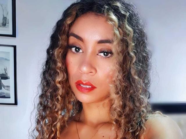 KylieJen with curly, blonde-highlighted hair, red lipstick, and brown eyes, looking directly at the camera, wearing a gold necklace. She's got a sexy, attractive face.